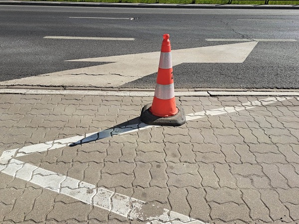 Pomarańczowy pachołek drogowy na utwardzonej powierzchni z białymi oznaczeniami parkingowymi.