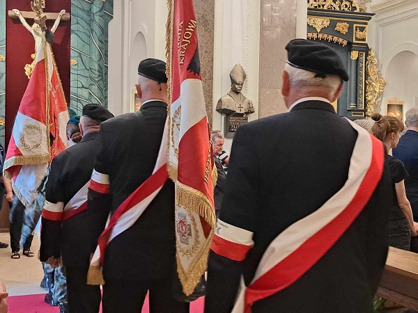 W ceremonii biorą udział ludzie ubrani w formalne stroje i przepasani polskimi szarfami wojskowymi.
