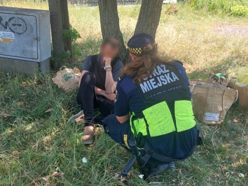 Policjant pomaga osobie siedzącej na ziemi w pobliżu jej rzeczy.