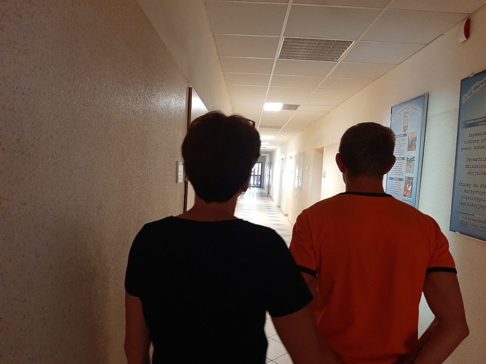 Dwie osoby idące korytarzem, jedna w czarnej koszulce, a druga w pomarańczowej.