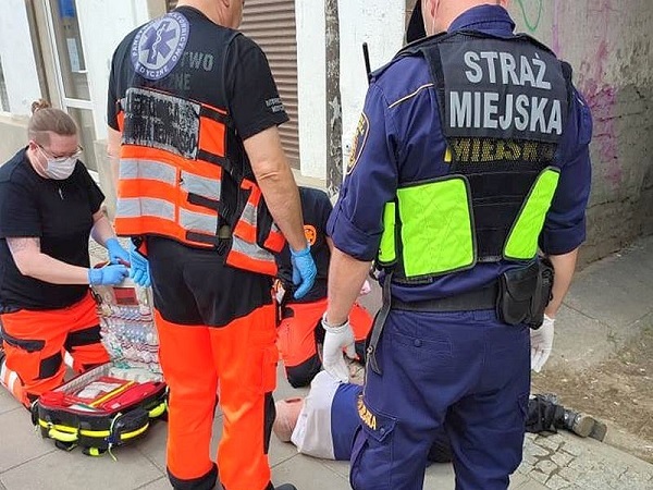Personel medyczny udzielający pomocy pacjentowi na ziemi. Na mundurze widoczny napis „Straż Miejska”.