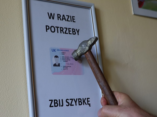 Hammer gotowy do wybicia szyby samochodu obok oprawionego brytyjskiego prawa jazdy. Tekst brzmi: „W razie potrzeby wybij szybę.