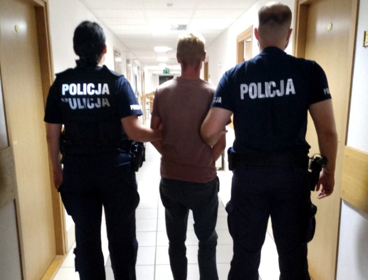 Dwóch polskich policjantów eskortuje podejrzanego korytarzem. Na ich mundurach widać napis POLICJA.