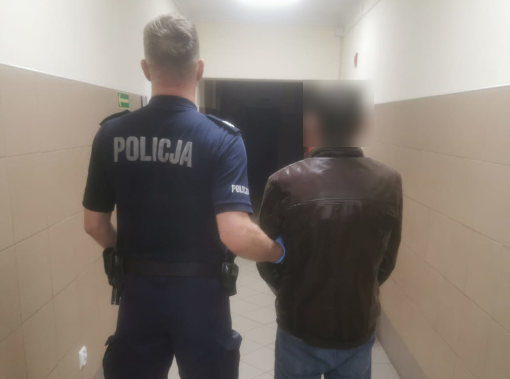Policjant aresztujący podejrzanego na korytarzu. Na mundurze policjanta widnieje napis „POLICJA.