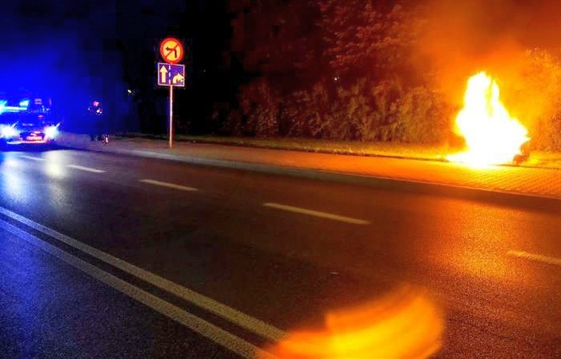 Nocny pożar samochodu w pobliżu znaku drogowego wskazującego zakręt. Widoczne są światła pojazdu uprzywilejowanego.