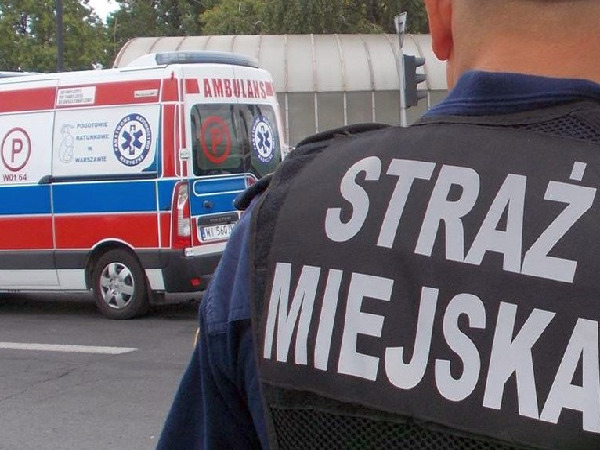 Plecy funkcjonariusza Straży Miejskiej, na mundurze dobrze widoczny „Straż Miejska”, w tle karetka.