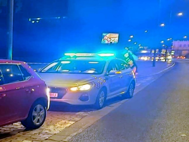 Samochód policyjny z włączonymi światłami zatrzymał się nocą w pobliżu różowego samochodu.