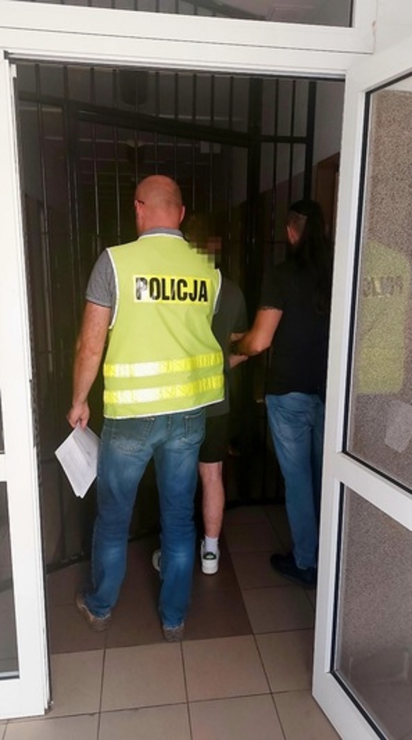Policjanci eskortujący podejrzanego do celi więziennej. Na kamizelce funkcjonariusza widać napis „POLICJA”.
