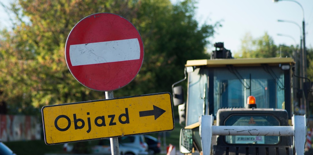 Znak drogi zamkniętej ze strzałką objazdu, wskazujący roboty drogowe z pojazdem budowlanym w tle. „Objazd” jest wyraźnie widoczny.