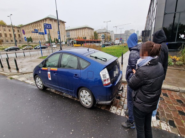 Niebieska taksówka Toyota Prius zatrzymała się na ulicy miejskiej, w pobliżu są ludzie.