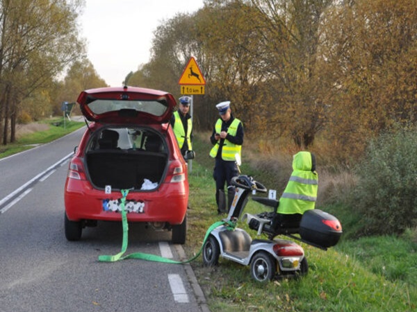 Policjanci badają wypadek skutera elektrycznego z udziałem czerwonego samochodu.