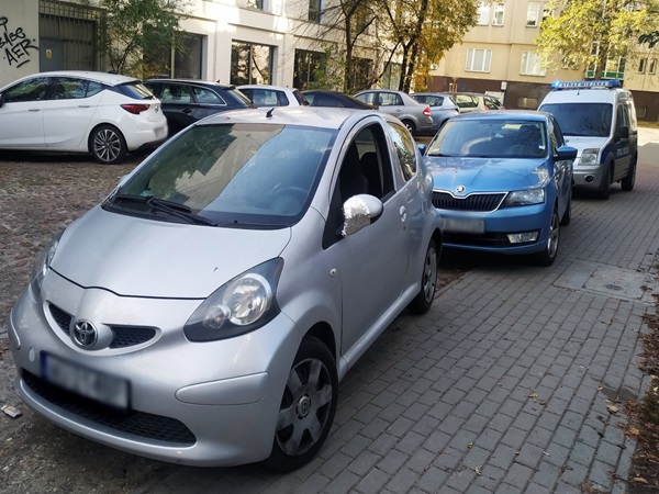 Srebrna Toyota Aygo zaparkowana nielegalnie obok niebieskiej Skody, z tyłu znajduje się policyjny furgon.