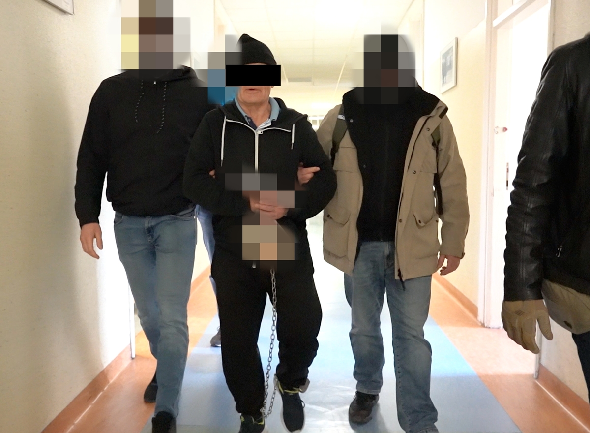 Więzień w kajdankach eskortowany przez strażników wzdłuż korytarza szpitalnego.