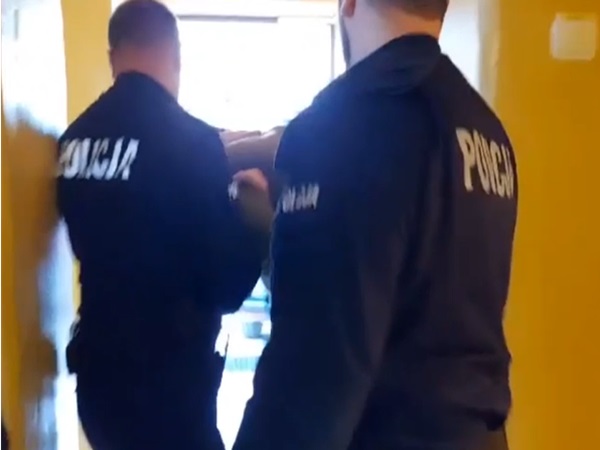 Dwóch policjantów eskortuje podejrzanego korytarzem. Na ich mundurach widać napis „POLICJA”.