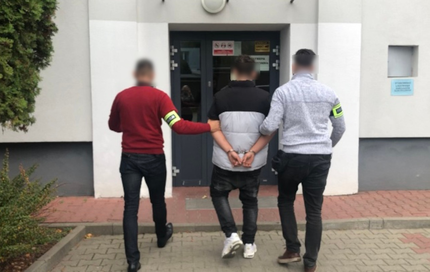 Dwóch funkcjonariuszy eskortuje skutego kajdankami podejrzanego z budynku.