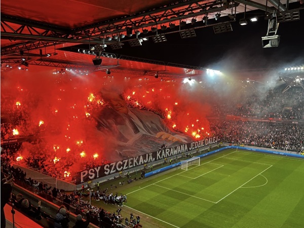 Czerwone flary i wyświetlacz tifo na stadionie piłkarskim, na którym widać duży baner z napisem „Psy Szczekaja, Karawana Jedzie Dalej.