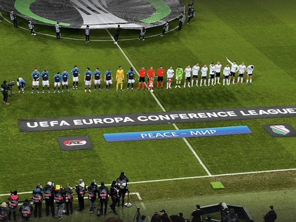 Mecz Ligi Konferencji Europejskiej UEFA: Dwie drużyny ustawiają się do rozpoczęcia meczu, wyświetlany jest baner z napisem „Peace - Мир”.