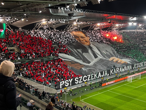 Ogromny tifo stadionu przedstawiający legendarnego menadżera Lecha Poznań, a kibice trzymają flagi i transparenty z napisem „Psy szczekają, karawana jedzie dalej”.