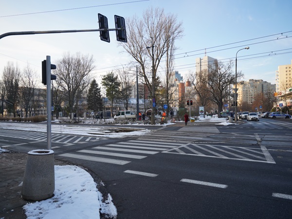 Zaśnieżone skrzyżowanie z przejściem dla pieszych, sygnalizacją świetlną i budynkami w tle.