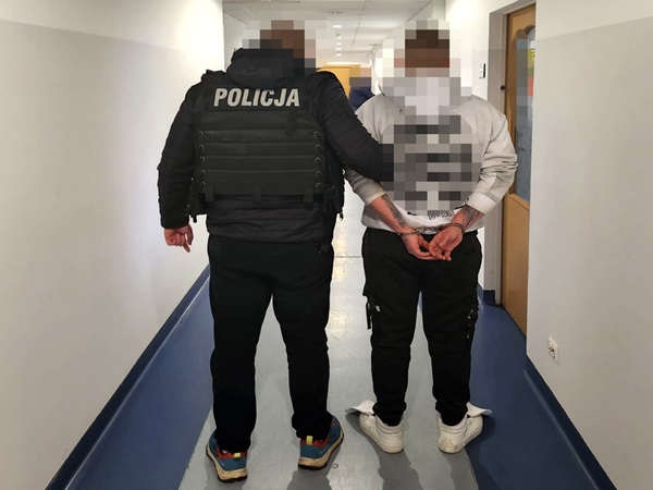 Policjant eskortujący podejrzanego w kajdankach korytarzem. Na kamizelce policjanta widoczny jest napis „POLICJA”.