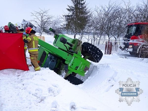 Strażacy ratują zielony traktor z zaśnieżonego rowu. Na miejscu jest policja.