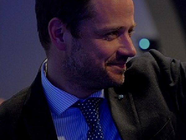 Zdjęcie mężczyzny w garniturze i niebieskiej koszuli w paski, uśmiechniętego.