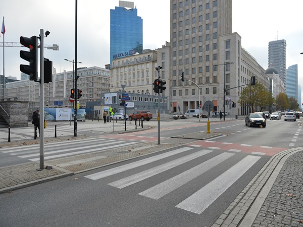 Scena uliczna Warszawy z przejściem dla pieszych, sygnalizacją świetlną i nowoczesnymi budynkami.