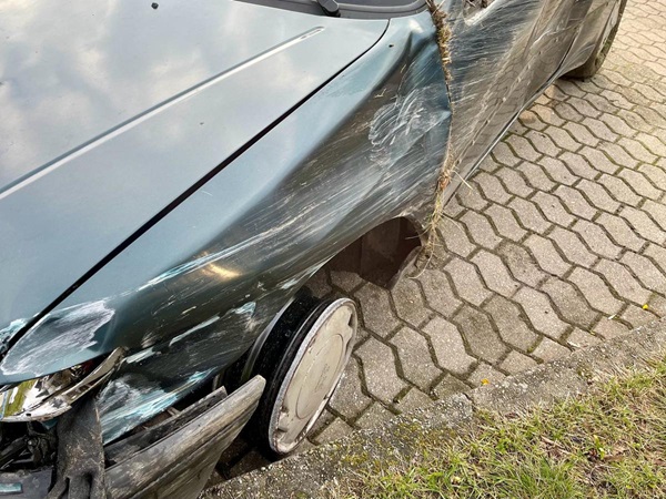 Uszkodzony ciemnozielony samochód z powodu kolizji czołowej, na karoserii widoczne są znaczne zarysowania i otarcia.