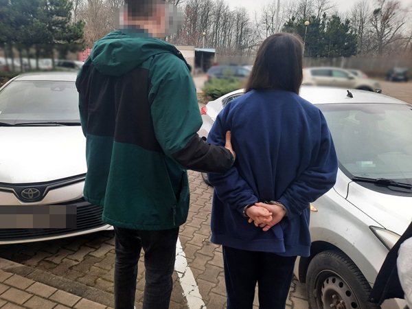 Policjant aresztujący skutą kajdankami kobietę przy zaparkowanych samochodach.