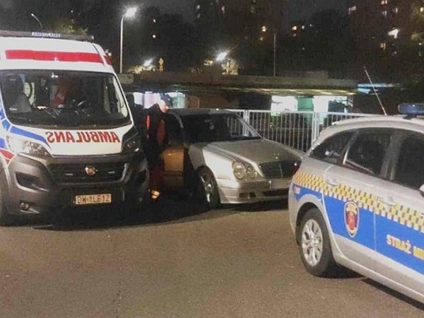 Ratownicy medyczni na nocnej scenie: karetka pogotowia, radiowóz i uszkodzony samochód.