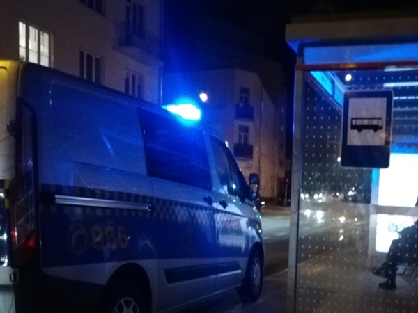 Policyjny furgon z niebieskimi światłami zaparkowany nocą w pobliżu przystanku autobusowego.