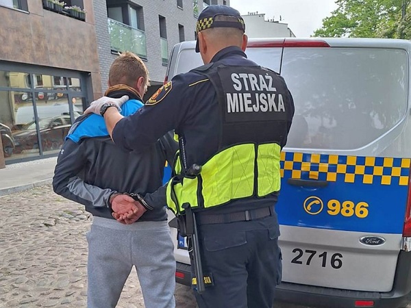 Policjant aresztujący podejrzanego, widoczne kajdanki. W tle pojazd Straży Miejskiej.