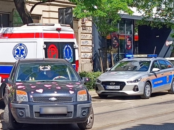 Samochód ślubny udekorowany różowymi sercami zaparkowany obok karetki pogotowia i radiowozu.