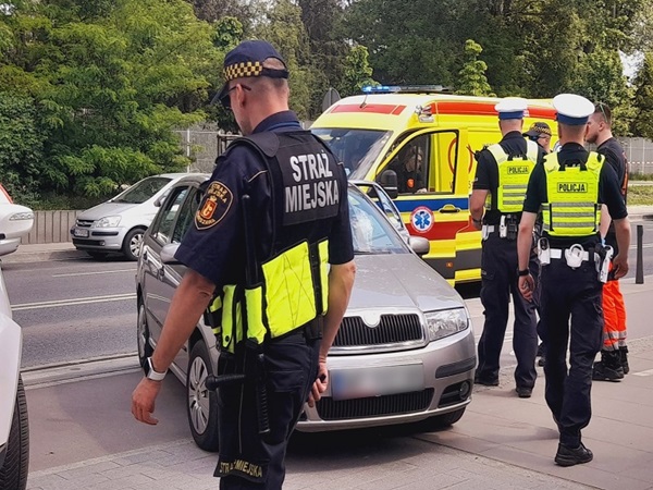 Polscy policjanci na miejscu wypadku drogowego, z widoczną karetką i uszkodzonymi samochodami. Na mundurze oficera widać napis „Straż Miejska”.