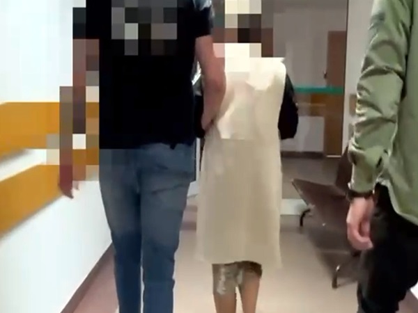 Dwie osoby eskortują osobę w szpitalnej koszuli wzdłuż korytarza.