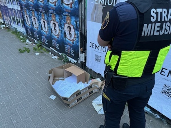Porzucone papiery w tekturowym pudełku na miejskim chodniku w pobliżu strażnika. Na mundurze strażnika widoczny napis „Straż Miejska”.