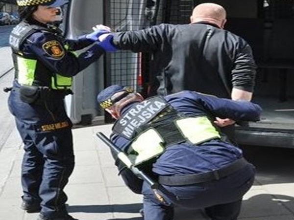 Trzech strażników miejskich zatrzymuje podejrzanego; jeden ze strażników zakłada mu kajdanki, a drugi obserwuje.