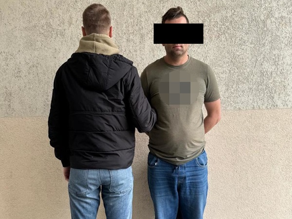 Dwóch mężczyzn stoi pod ścianą, jeden w czarnej kurtce, a drugi w zielonej koszuli. Aresztowania.