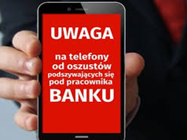 Ręka trzymająca smartfon wyświetlający ostrzeżenie o oszustwach związanych z telefonami bankowymi. Tekst brzmi: UWAGA na telefony od oszustów podszywających się pod pracownikiem BANKU.