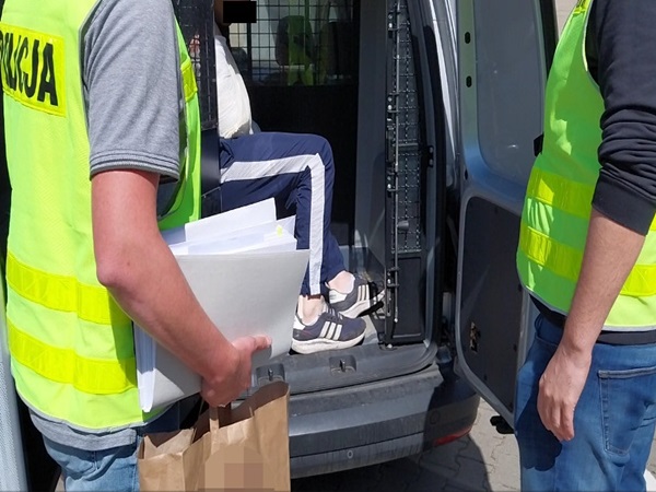 Policjanci eskortują podejrzanego do furgonetki, trzymając w rękach dokumenty.