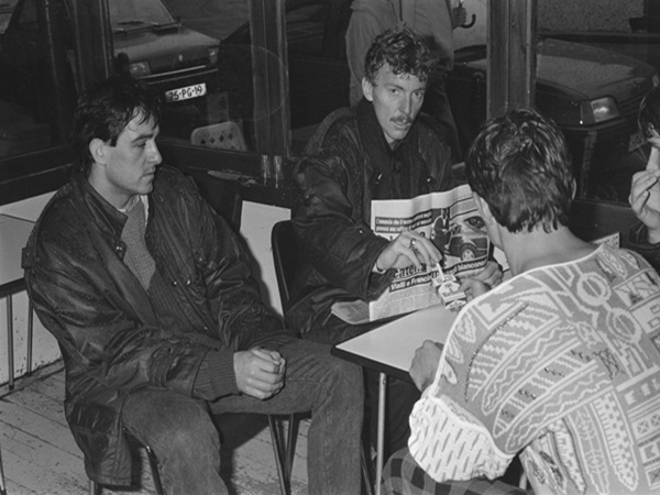 Trzech młodych mężczyzn siedzi przy stole, pali i czyta gazetę.