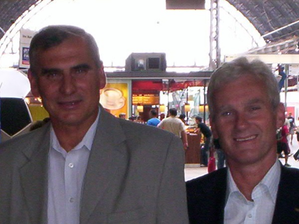 Dwóch mężczyzn, prawdopodobnie koledzy lub przyjaciele, pozują do zdjęcia na dworcu kolejowym.