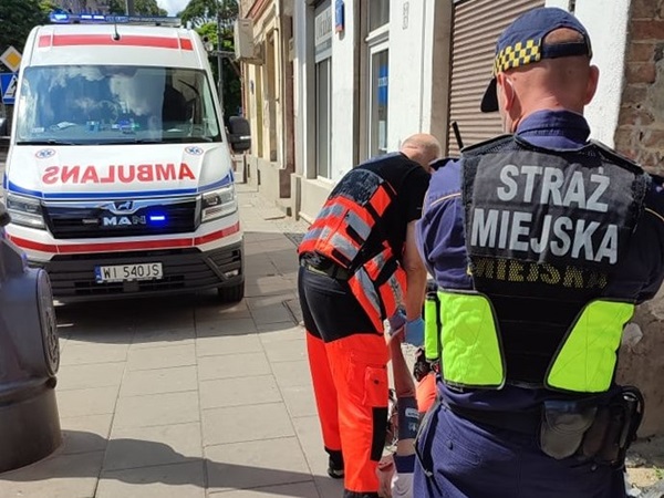 Ratownicy medyczni pomagają osobie znajdującej się w pobliżu karetki pogotowia. Na mundurze widoczna jest Straż Miejska.