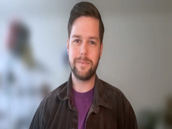 Zdjęcie mężczyzny z brodą, ubranego w ciemną kurtkę i fioletową koszulę.