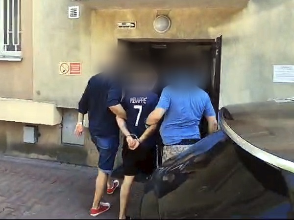 Policjanci aresztujący podejrzanego w kajdankach przed budynkiem. Na koszulce podejrzanego widoczny jest napis „MBAPPE 7”.