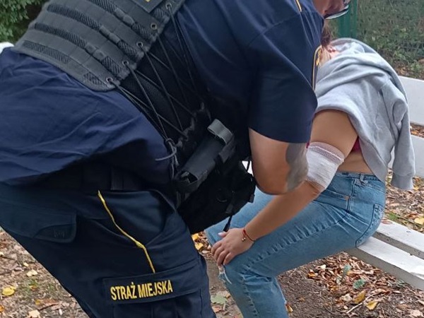 Strażnik miejski pomaga rannej kobiecie na ławce w parku.