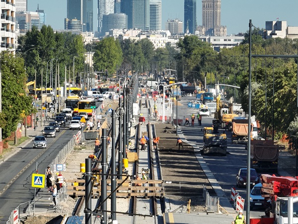 Budowa linii tramwajowej w Warszawie. Na zdjęciu widać robotników i ciężki sprzęt na ruchliwej ulicy miasta.