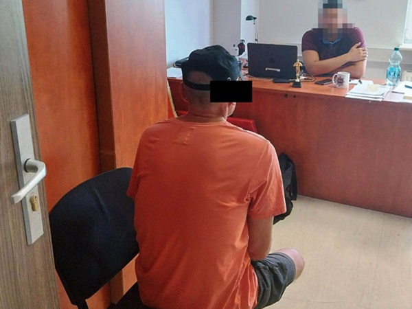 Mężczyzna w pomarańczowej koszuli siedzi podczas przesłuchania przez policję. Funkcjonariusz siedzi po drugiej stronie biurka.