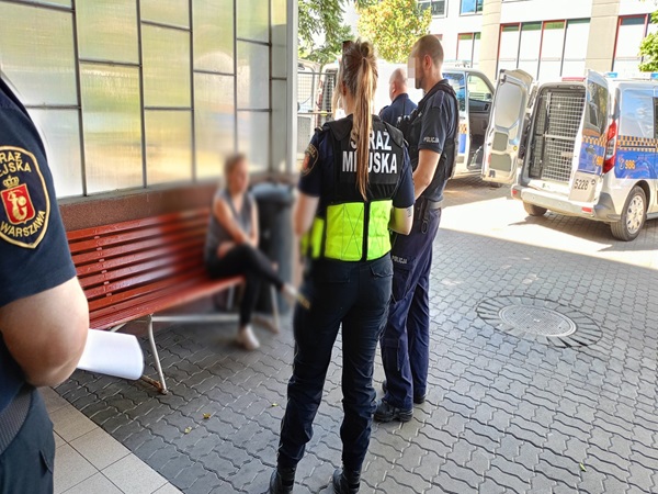 Policjanci zatrzymują kobietę na ławce obok radiowozu.
