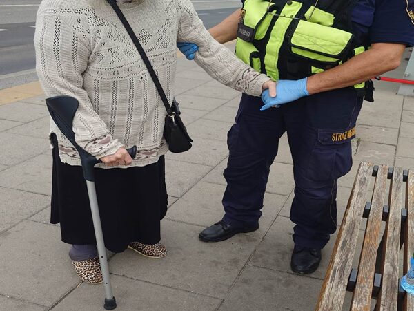 Starsza kobieta z laską, wspomagana przez umundurowanego policjanta.